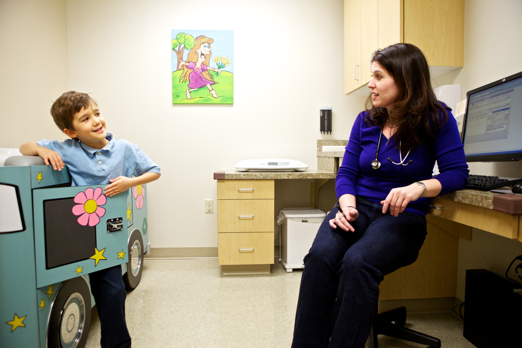 A young boy smiles and talks to a female doctor in a medical office. The boy stands by a play structure shaped like a bus, while the doctor sits near a computer. A colorful painting hangs on the wall behind them.