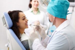 A dentist wearing a mask and gloves examines a smiling woman’s teeth in a dental chair, while another person in a white coat stands in the background observing.