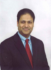 A man wearing a dark suit, light blue shirt, and red tie poses for a formal portrait against a plain white background. He has short dark hair and is smiling slightly at the camera.