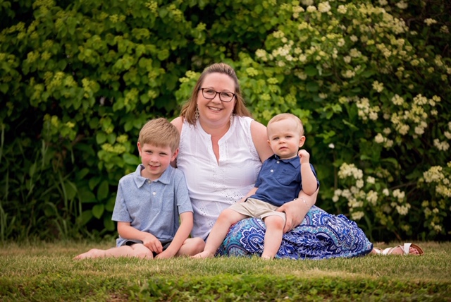 A woman sits on the grass in front of green bushes, smiling with two young boys beside her. One boy sits on her lap, while the other sits close to her side. All three look happy and relaxed outdoors.