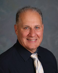 Smiling older man with short gray hair wearing a dark dress shirt and a light-colored striped tie, posing in front of a neutral, gray-blue background.