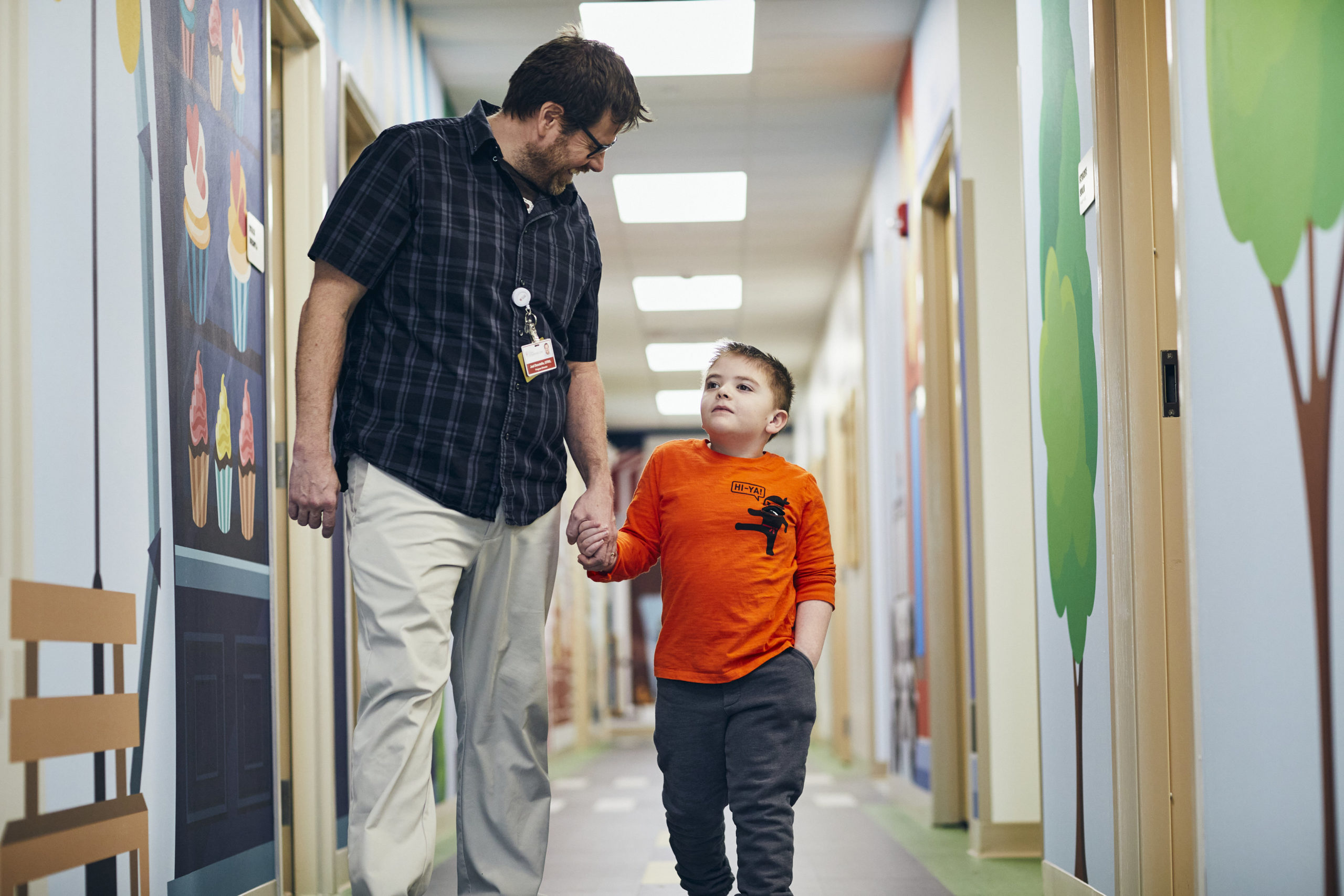 An adult and a young boy walk hand-in-hand down a brightly decorated hallway. The boy wears an orange shirt and looks up at the adult, who is smiling and wearing glasses, a name badge, and a checkered shirt.