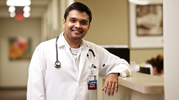 A smiling male doctor wearing a white coat and stethoscope stands at a reception desk in a medical office. The background is softly blurred, showing a hallway and framed artwork on the wall.