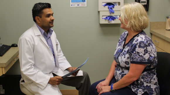 A doctor in a white coat sits and talks with a woman in medical scrubs in a clinic room; medical gloves and equipment are visible on the wall in the background.