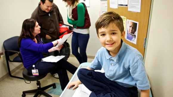 A young boy sits and smiles on an exam table in a doctor's office. Behind him, a woman talks to a seated healthcare worker while another adult stands nearby, holding a red bag.