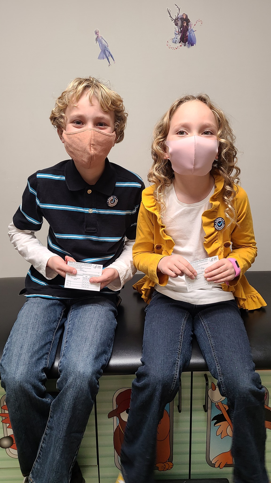 Two children wearing masks sit side by side on a bench, each holding a card. They both have light hair and wear casual clothes—a striped polo and jeans, and a yellow jacket with jeans. Cartoon and Frozen stickers are on the wall behind them.