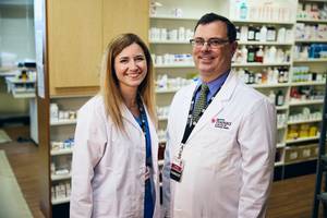 Two pharmacists wearing white lab coats stand and smile in a pharmacy, with shelves of medication and supplies visible in the background.