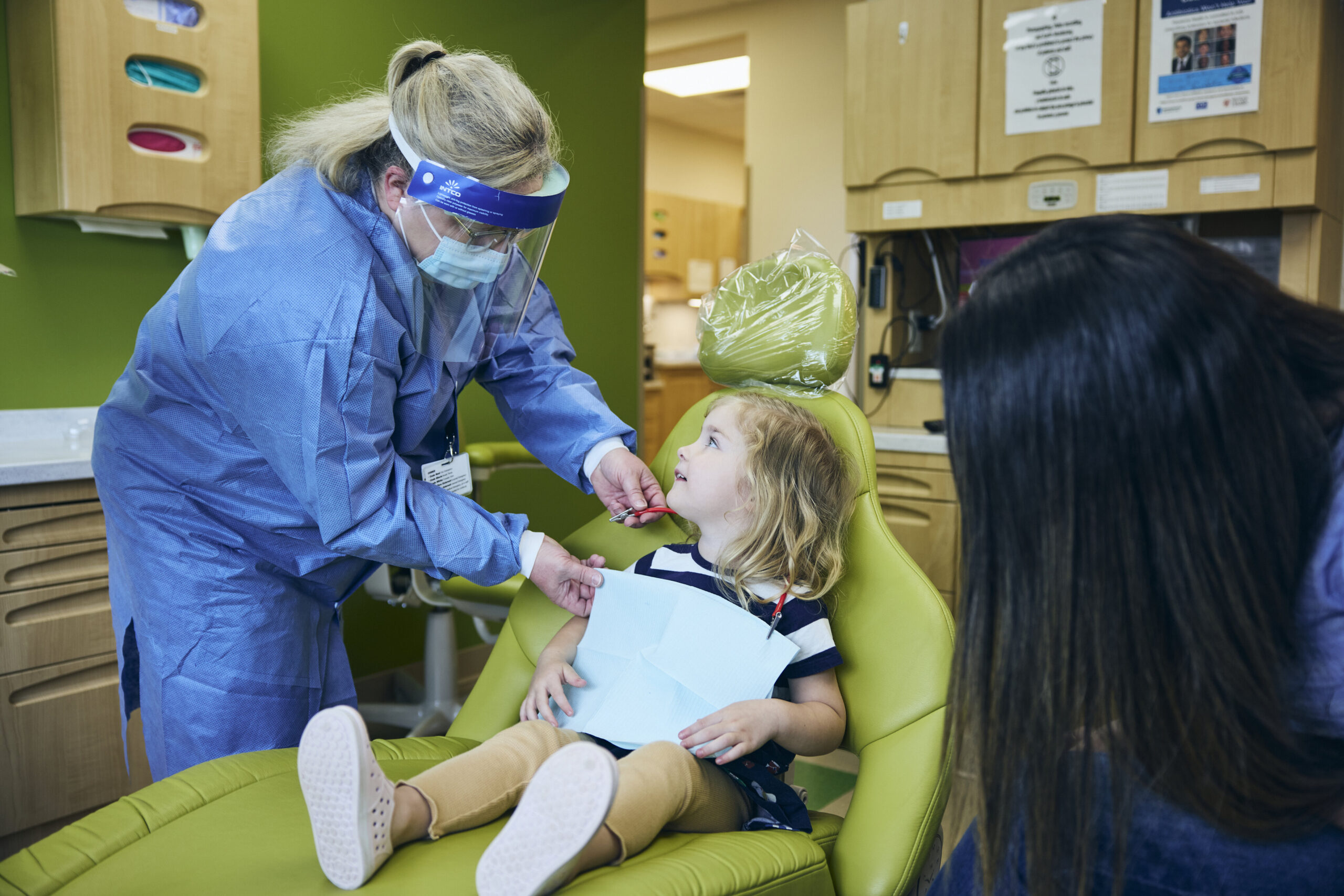 A dental professional in protective gear speaks to a young girl sitting in a dental chair, while an adult sits nearby. The setting is a brightly lit dentist’s office with green walls and wooden cabinets.