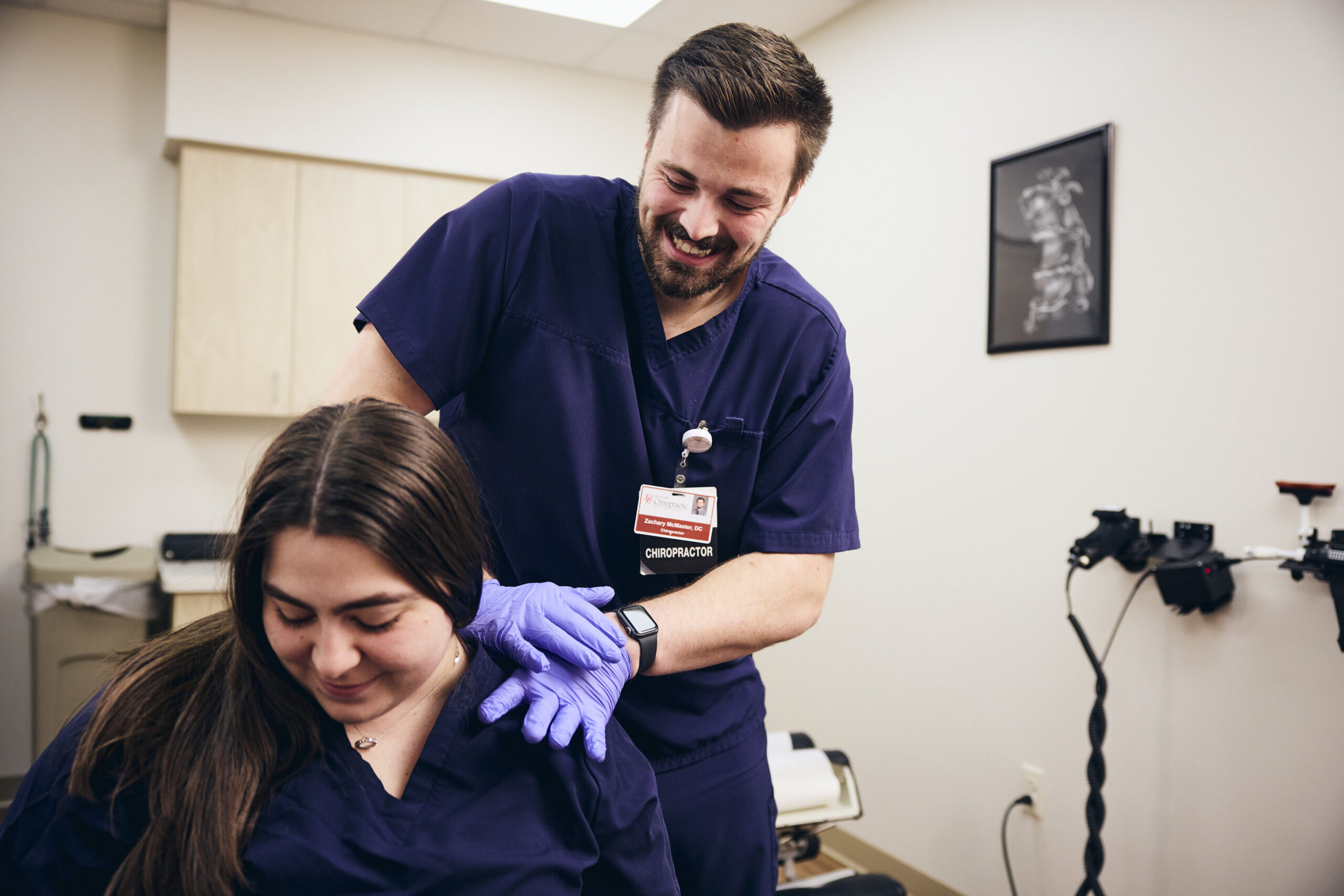 A smiling healthcare professional in navy scrubs and gloves gently adjusts a seated woman's shoulder in a medical office. Both appear relaxed, and medical equipment is visible in the background.