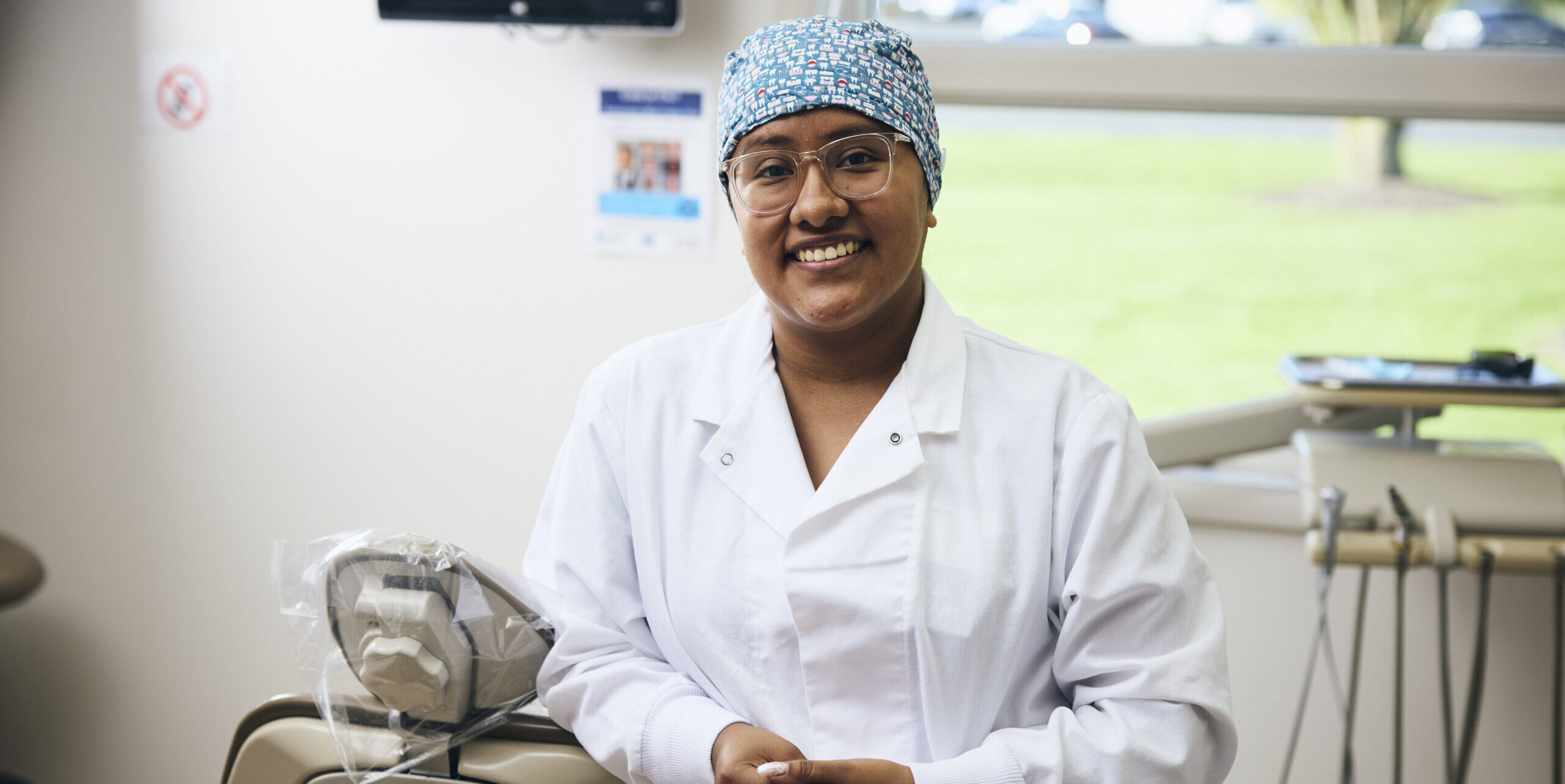A smiling healthcare worker in a white coat and patterned surgical cap stands in a bright medical or dental office, with medical equipment visible in the background.