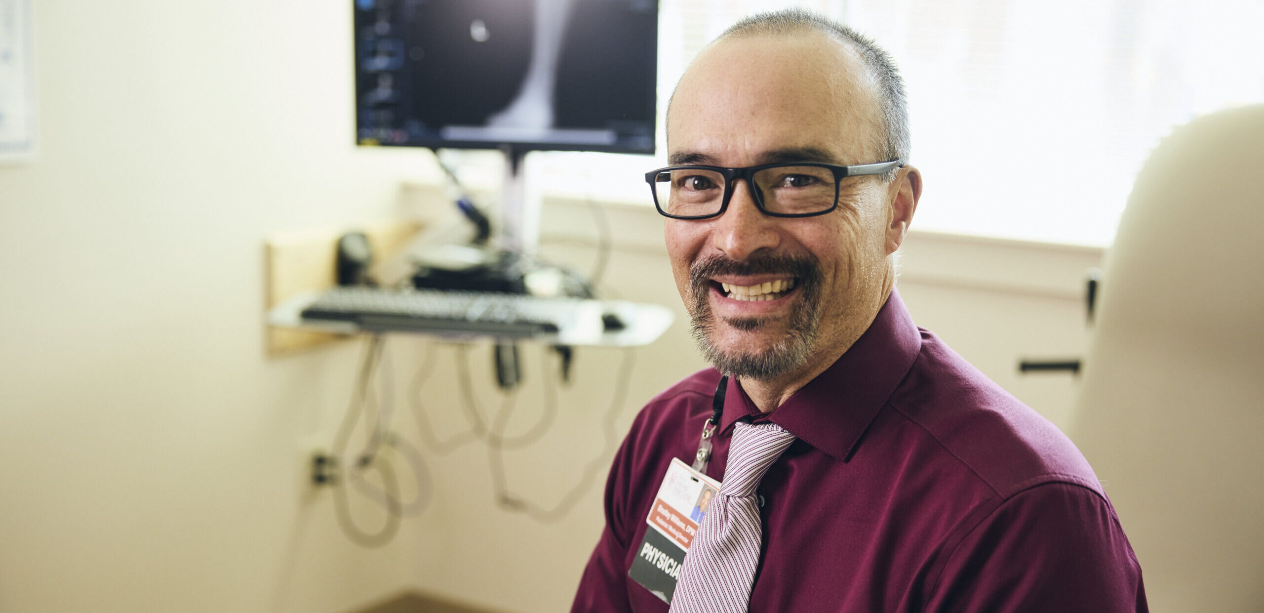 A man with glasses and a maroon shirt smiles at the camera while sitting in a medical office. A computer with an X-ray image is visible in the background.