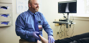 A medical professional wearing gloves examines a patient’s foot in a clinic. A computer monitor displaying an X-ray image is visible in the background.