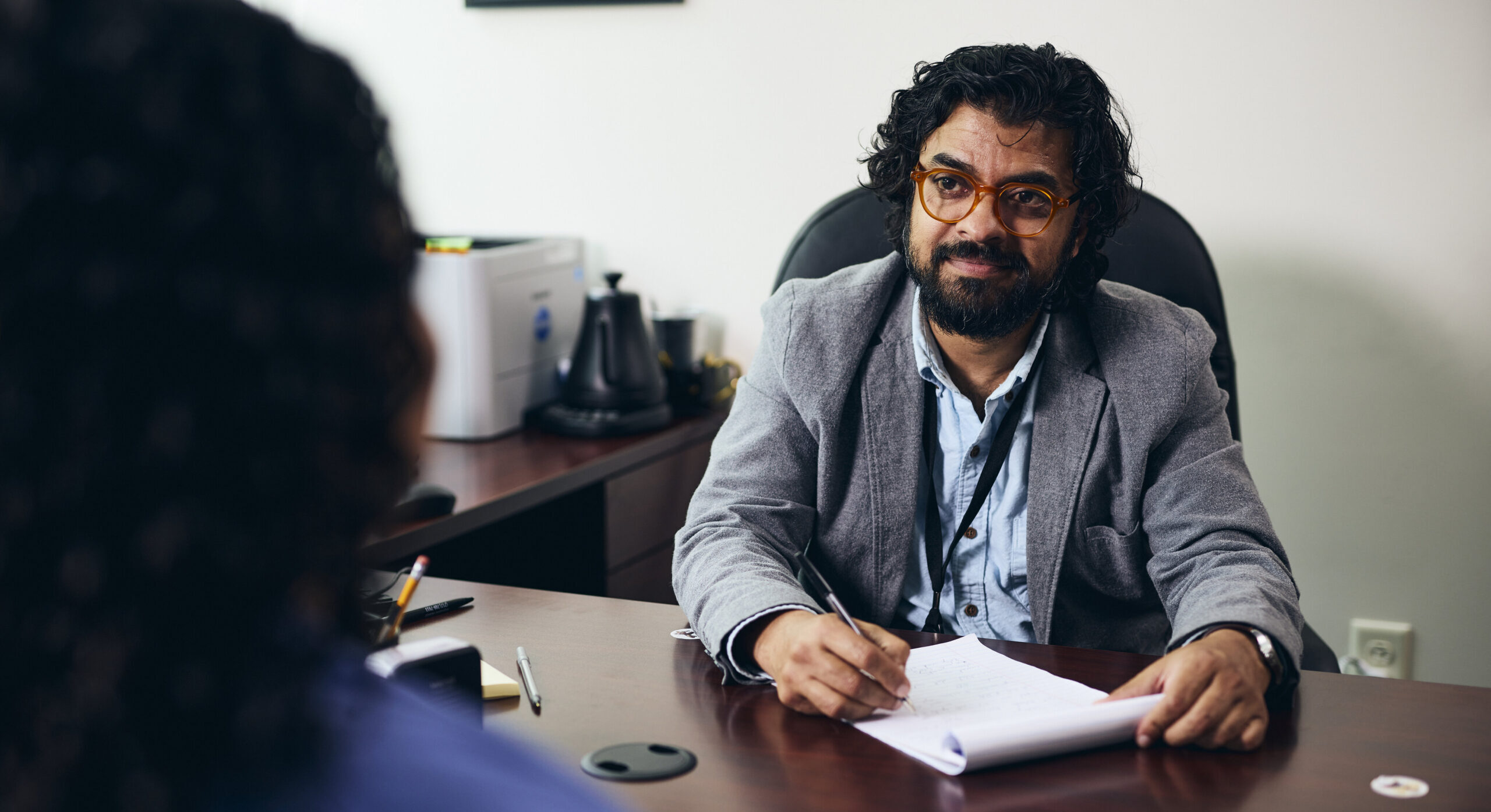 A man with curly hair, glasses, and a beard sits at a desk, holding a pen and paper, attentively listening to another person who is partially visible in the foreground.