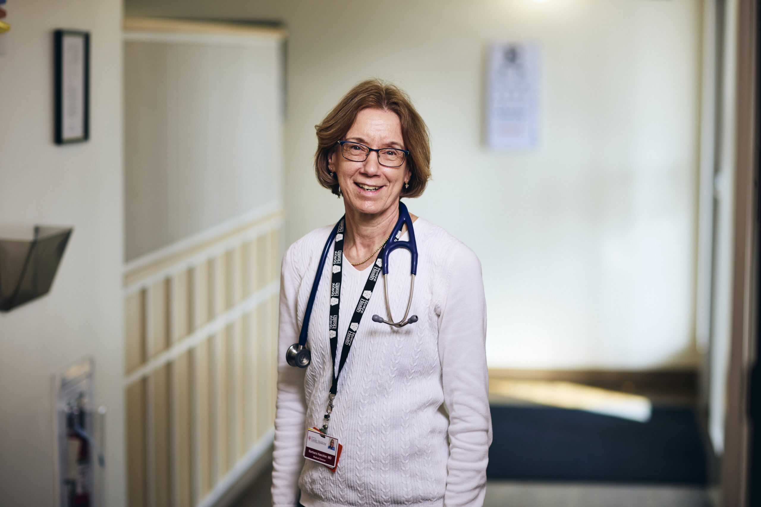 A smiling doctor wearing glasses, a white sweater, and a stethoscope around her neck stands in a well-lit hallway with medical charts and a blurred eye chart in the background.