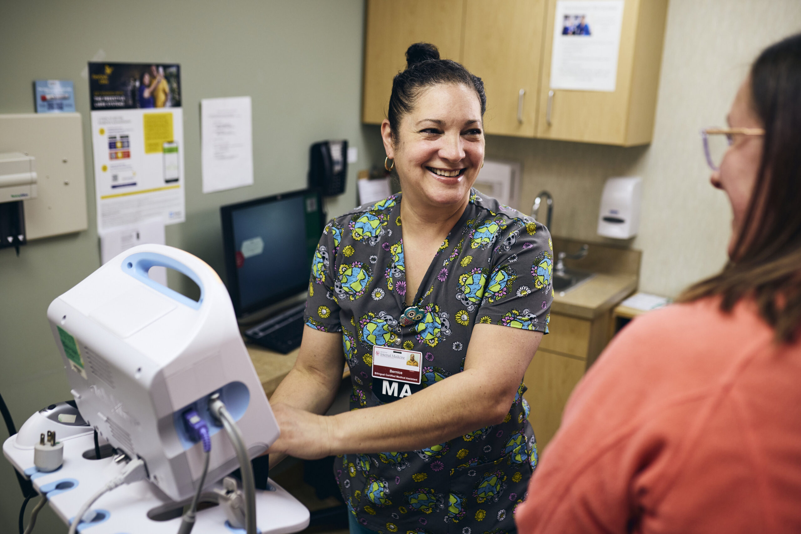 A smiling medical assistant in patterned scrubs stands next to medical equipment in an exam room, interacting with a patient wearing glasses and an orange top. The atmosphere appears friendly and professional.