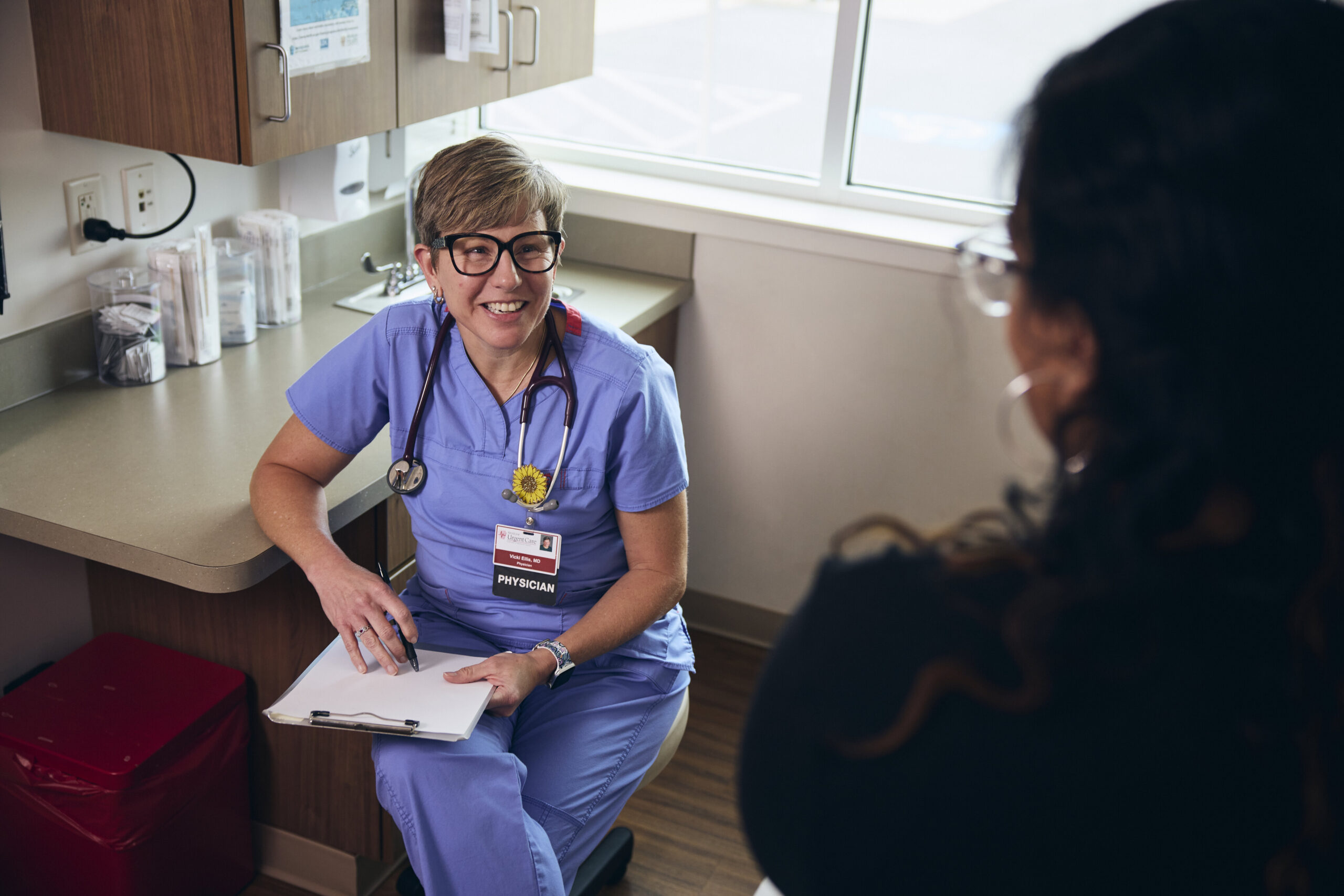 A smiling healthcare professional in scrubs and a stethoscope sits in a medical office, holding a clipboard and speaking with a patient seated across from them.