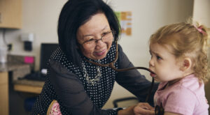 A smiling doctor uses a stethoscope to listen to a young girl’s chest during a checkup. The girl, wearing a pink shirt, looks curious and holds part of the stethoscope in her mouth.