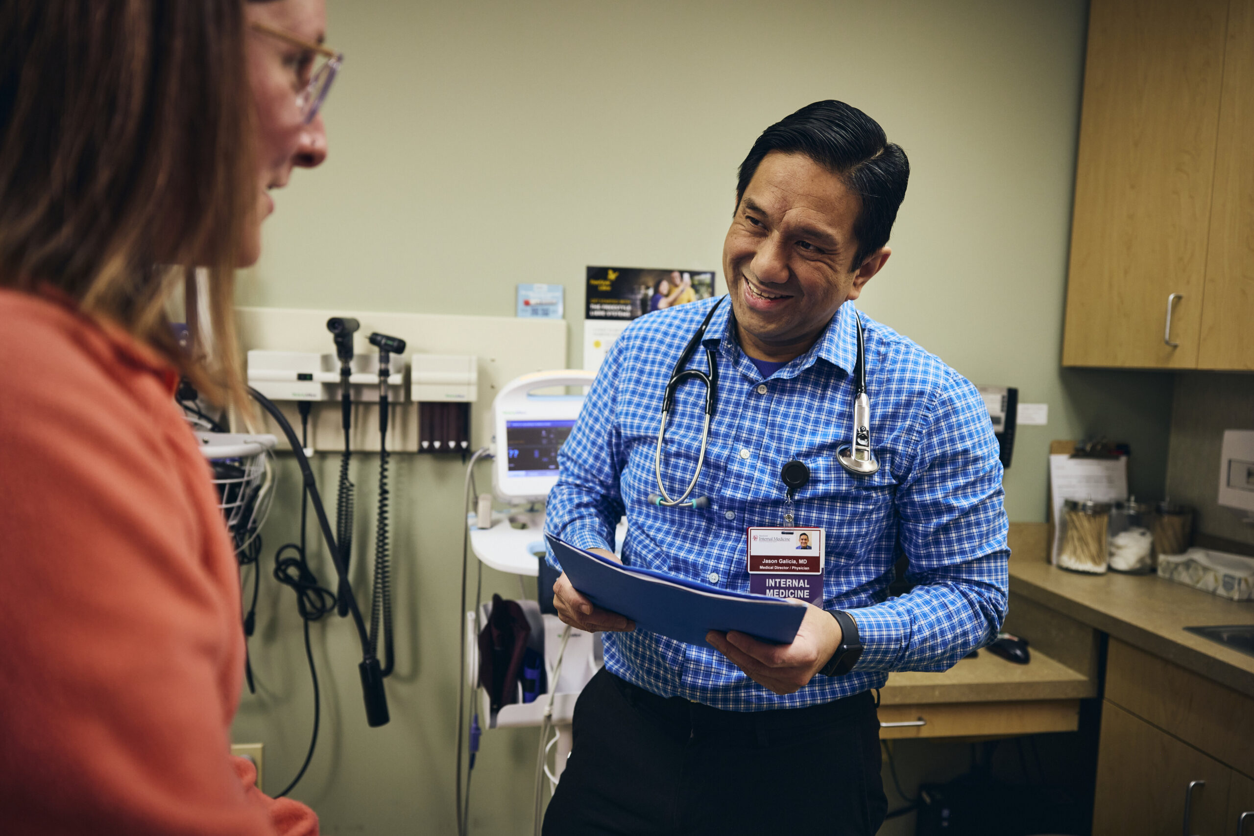 A doctor in a blue checkered shirt smiles while holding a clipboard and talking to a patient in a medical examination room with medical equipment in the background.