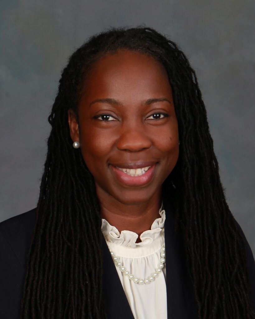 A woman with long braided hair, wearing a dark blazer, cream blouse with ruffles, and a pearl necklace, smiles in a professional headshot against a gray background.